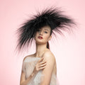 Woman wearing a large black feathered headpiece against a pink background