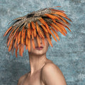Woman wearing a large, ornate hat with orange feathers against a textured gray background